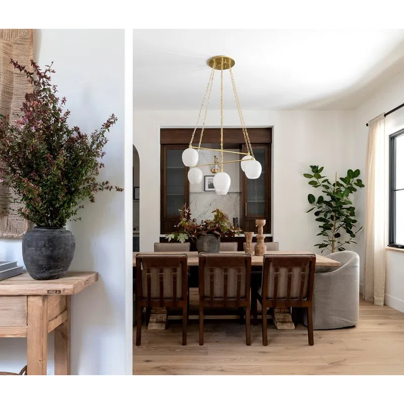 Bright modern dining room with wooden chairs, light hardwood floor, gold chandelier, and large green plants.