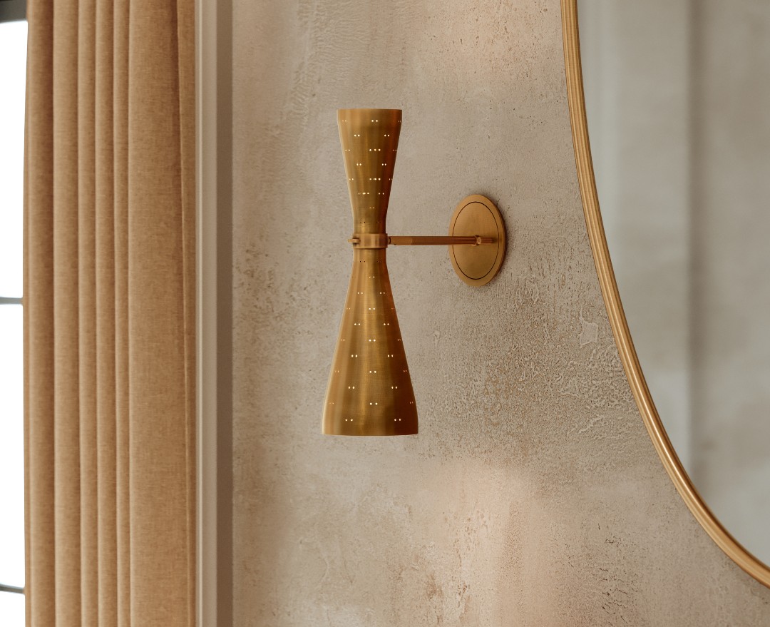 Modern brass wall sconce with perforated design mounted on textured beige wall beside large curved mirror.