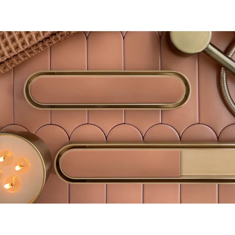 Modern pink bathroom accessories including soap dish, candle, brown towel, and brass showerhead on tiled surface.