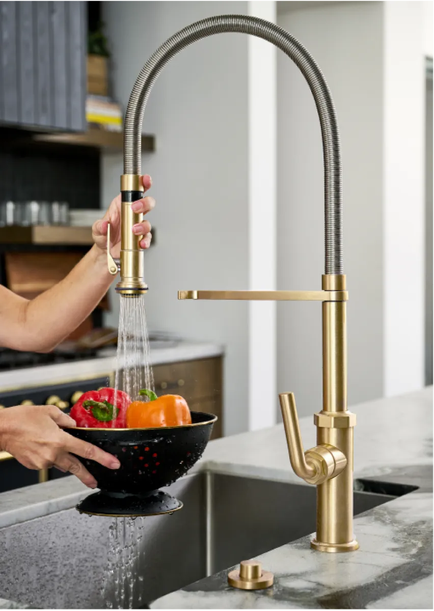 A brushed gold California Faucets pull-down kitchen faucet rinsing peppers in a colander over a stainless sink.