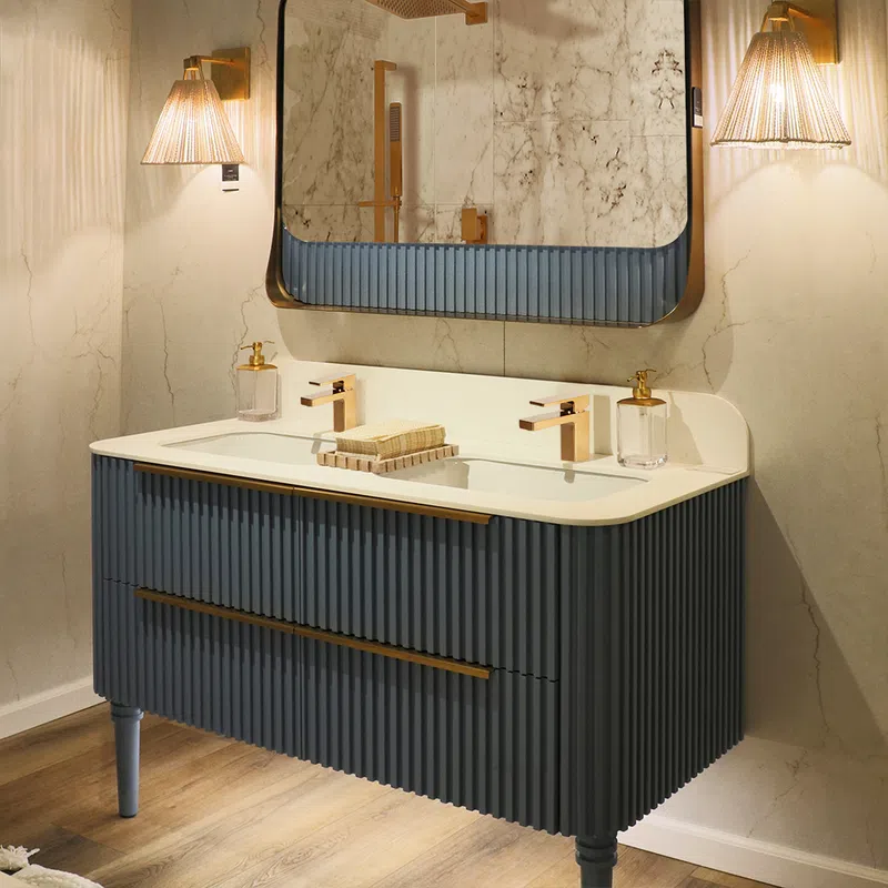 Modern bathroom vanity with ribbed dark blue cabinet, white countertop, dual gold faucets, and large mirror with lighting.