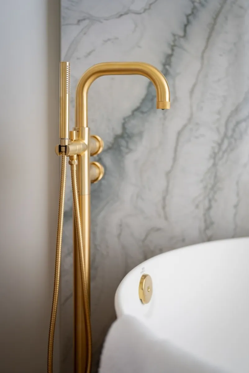 Brushed gold GRAFF freestanding bathtub faucet with handheld shower against a gray marble wall.