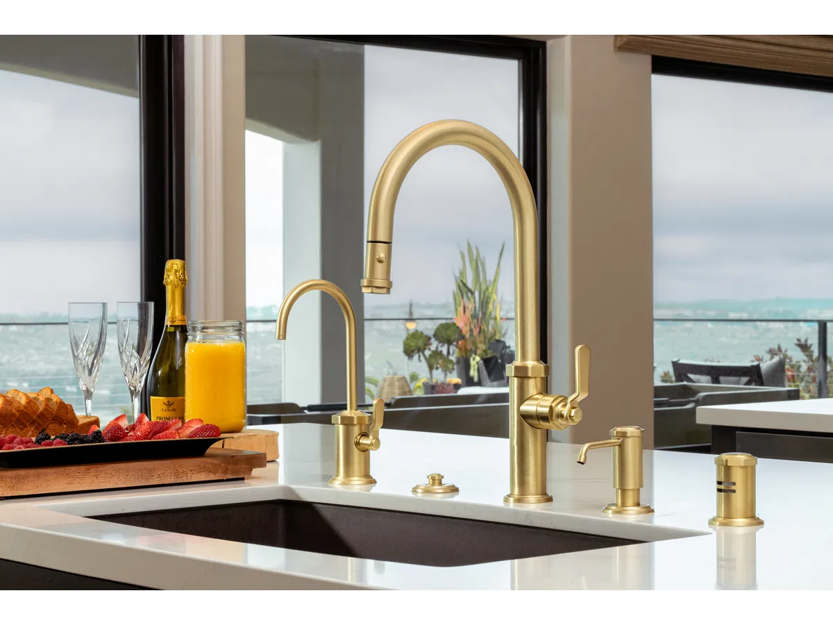 Modern kitchen sink with gold faucets and soap dispenser overlooking ocean view through large windows.