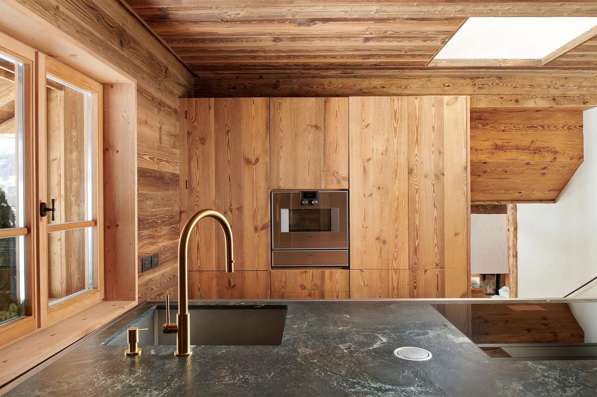 Modern kitchen with wooden walls, brass faucet, black marble countertop, built-in oven, and large window.