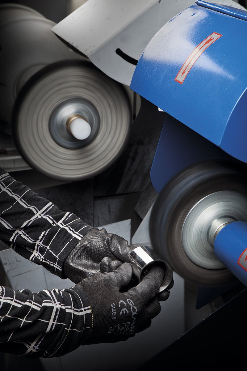 Worker wearing gloves polishing a metal ring using an industrial grinding machine with spinning wheels.