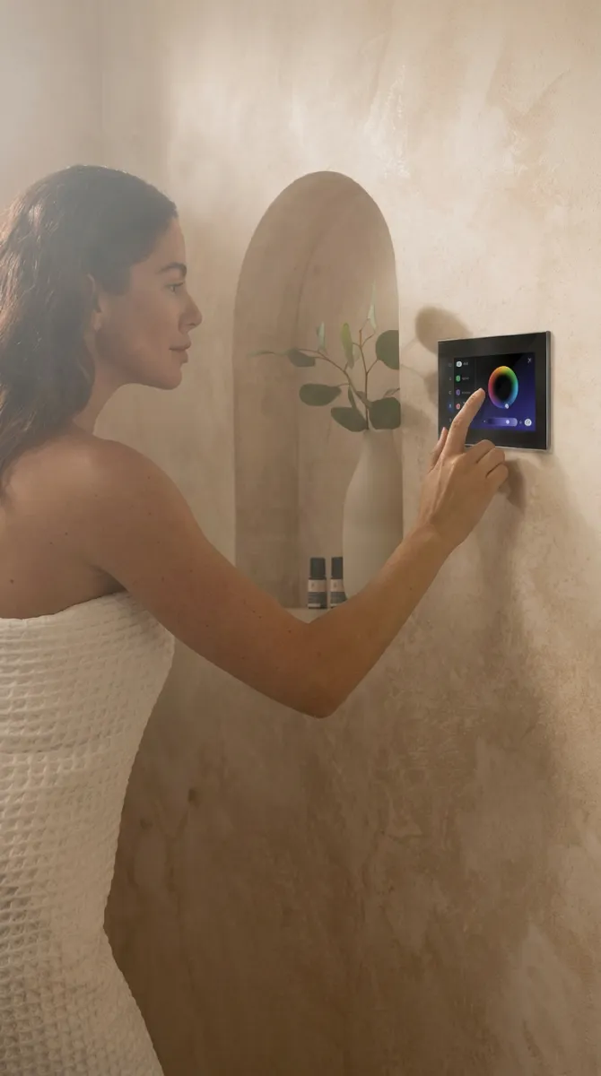Woman in a towel adjusting color settings on a wall-mounted touchscreen in a neutral-toned bathroom.