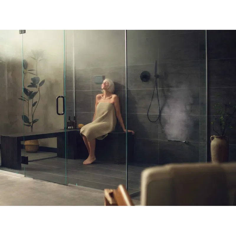 Woman wrapped in towel relaxing inside a modern glass steam shower with dark tiles and indoor plants.