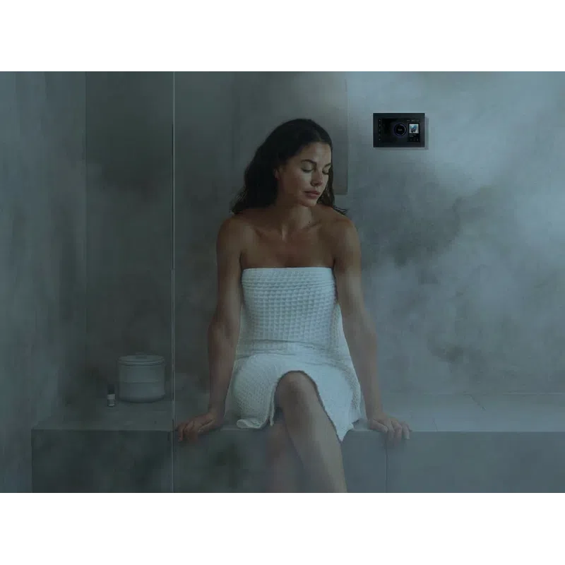 Woman in white towel relaxing on a steam room bench with a control panel on the wall amid steam.