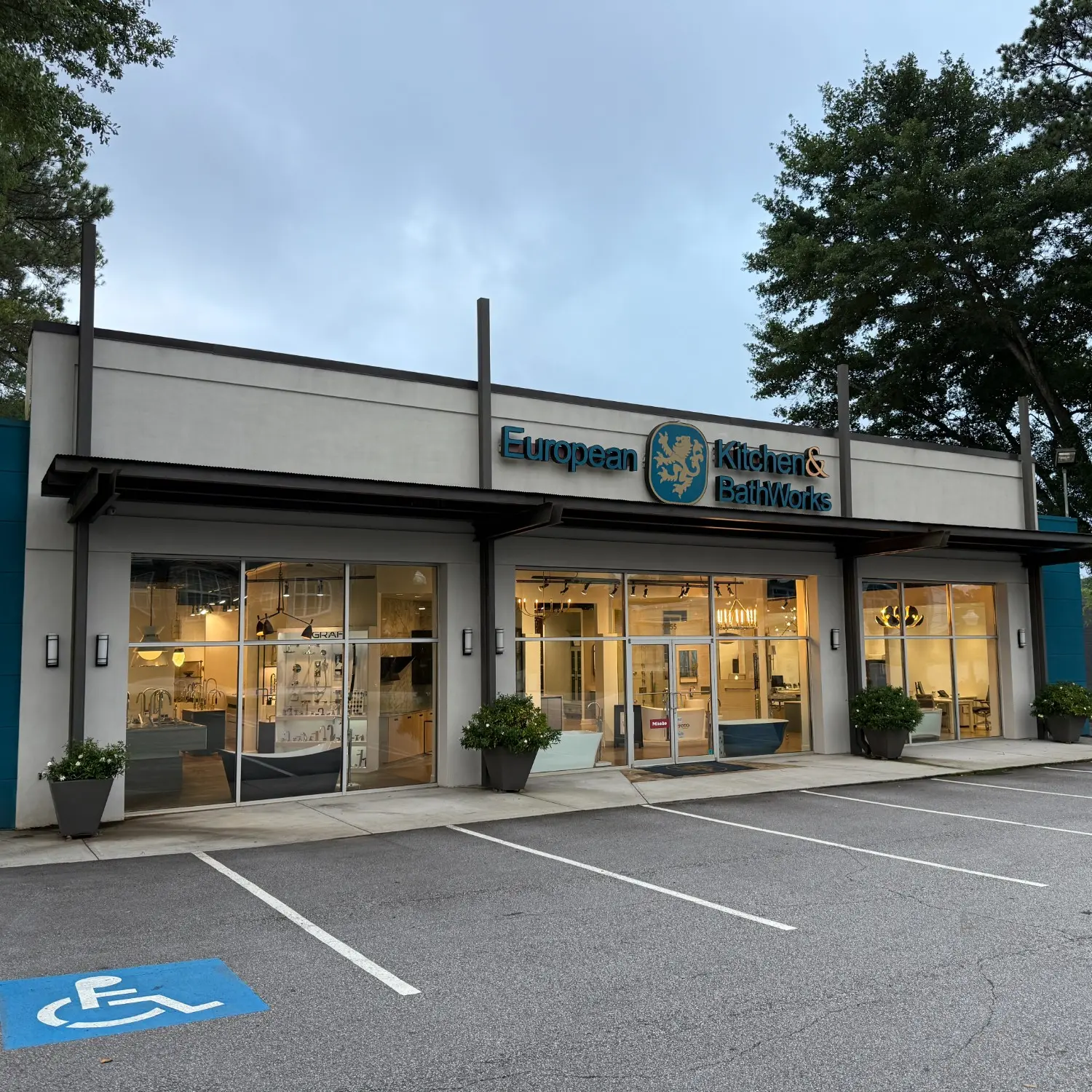 Exterior of European Kitchen & Bath Works showroom with glass windows, potted plants, and empty parking spaces at dusk.