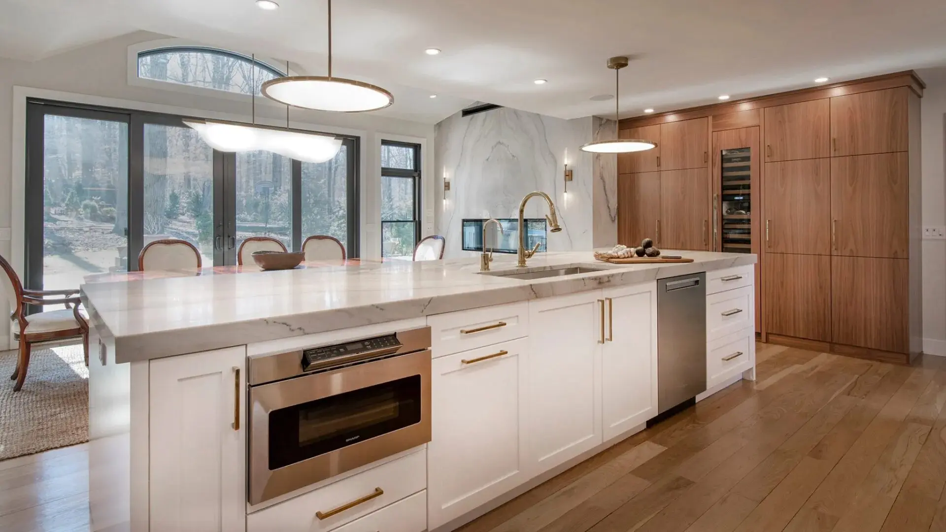 Modern kitchen with white marble island, wooden cabinets, stainless steel appliances, and large windows overlooking outdoor trees.