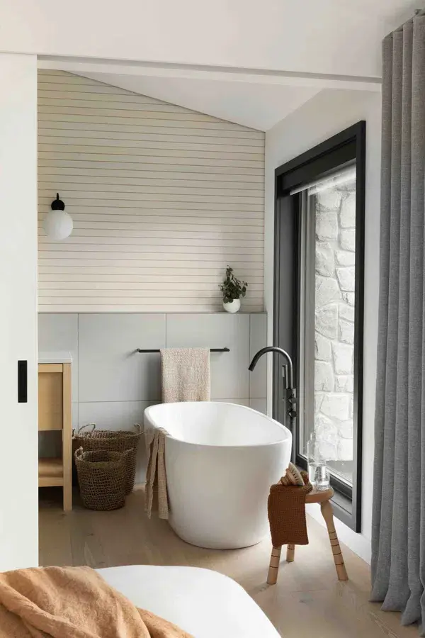 Freestanding white bathtub with a matte black Crosswater floor-mounted faucet in a minimalist bathroom with a large window.