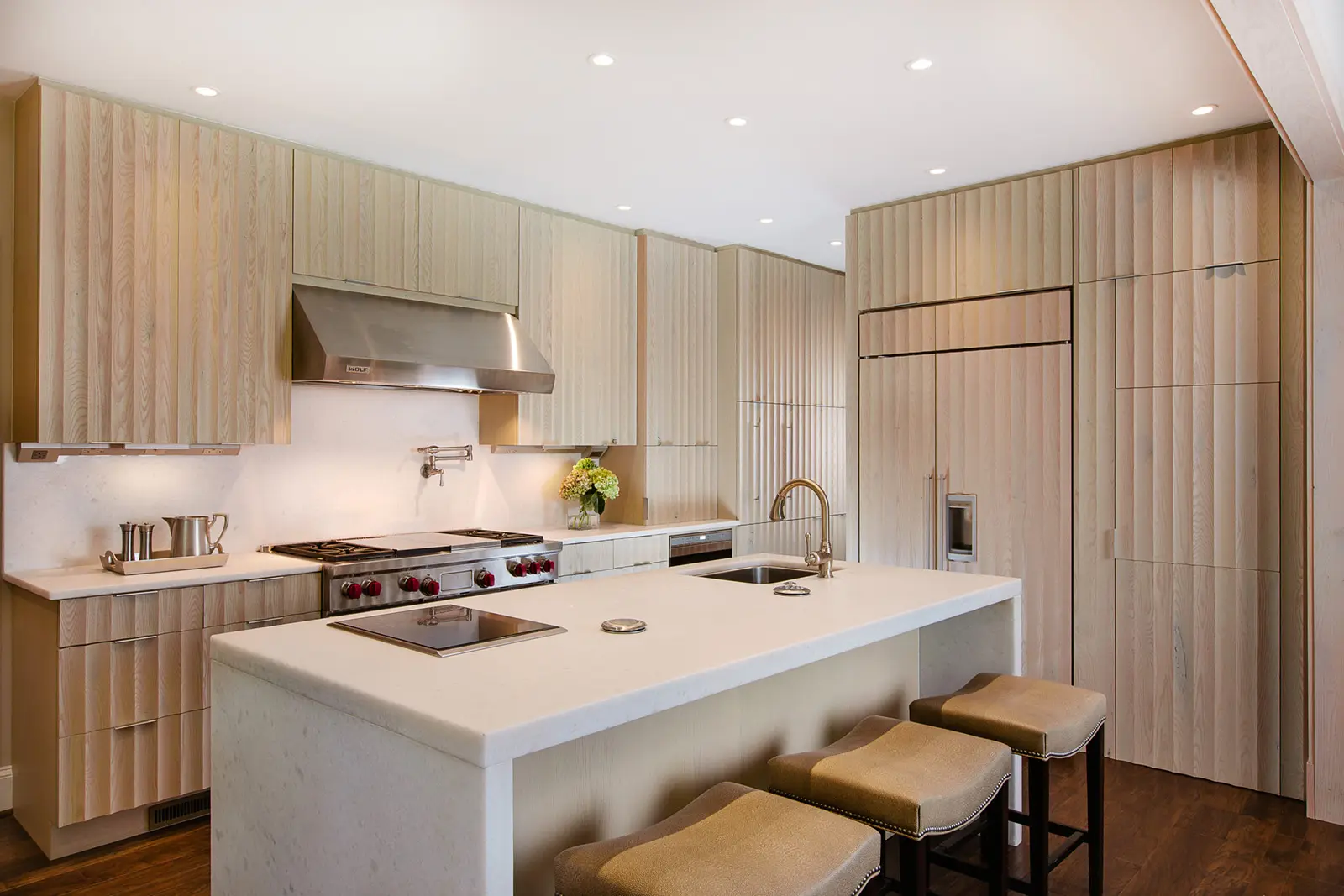 Modern kitchen with wooden cabinets, large white island with sink and cooktop, stainless steel hood, and bar stools.
