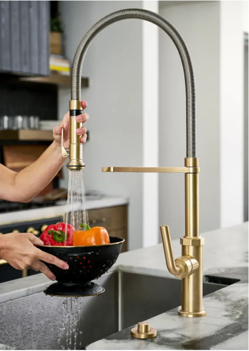 A brushed gold California Faucets pull-down kitchen faucet rinsing peppers in a colander over a stainless sink.