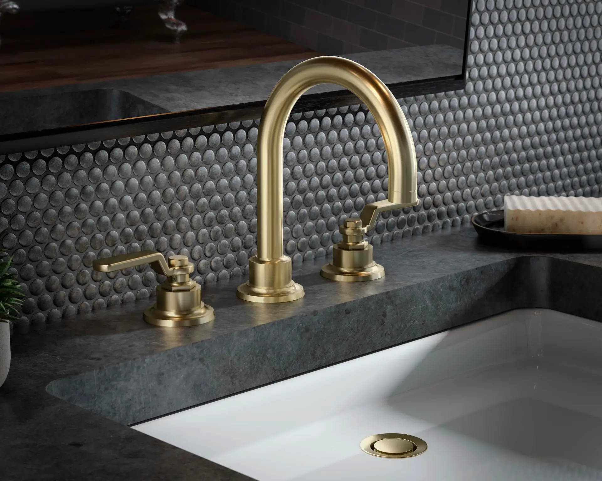 Brushed gold California Faucets widespread bathroom faucet on a dark stone vanity with penny-round tile backsplash.