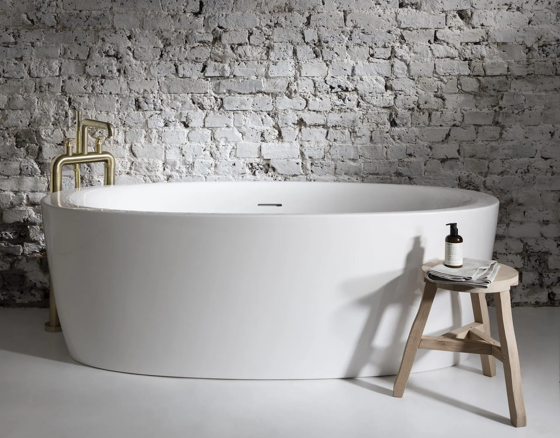 White freestanding soaking tub with a gold floor-mounted faucet against a whitewashed exposed brick wall.