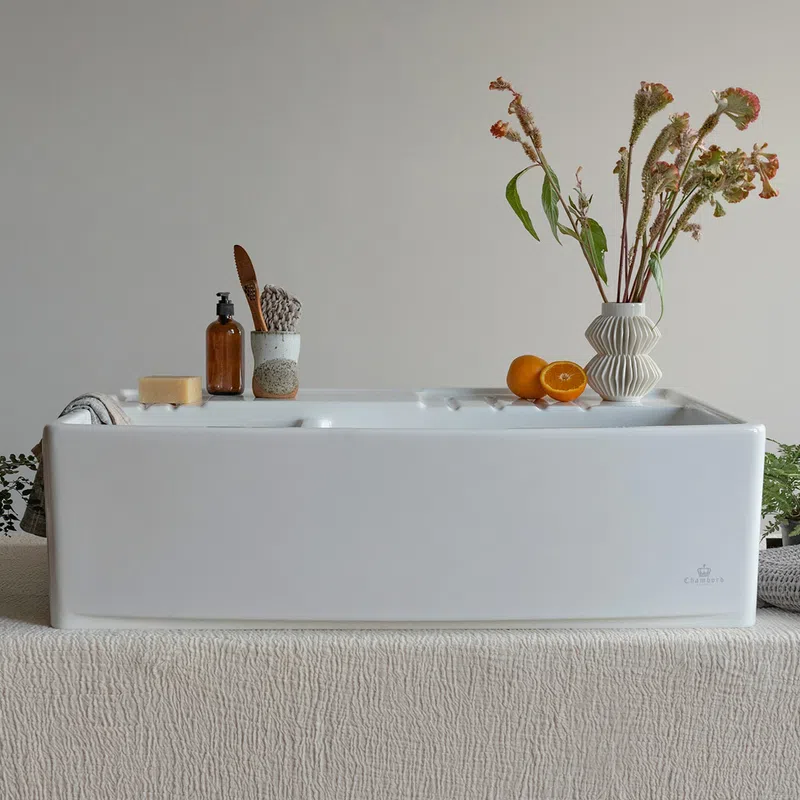 Modern white ceramic farmhouse sink on textured beige cloth with soap, brush, oranges, and vase with dried flowers.
