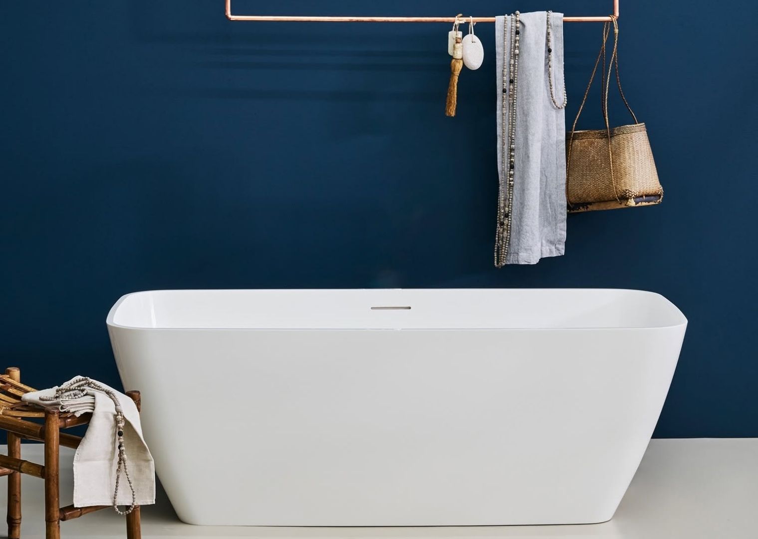 Modern white freestanding bathtub against dark blue wall with hanging towel and basket accessories.