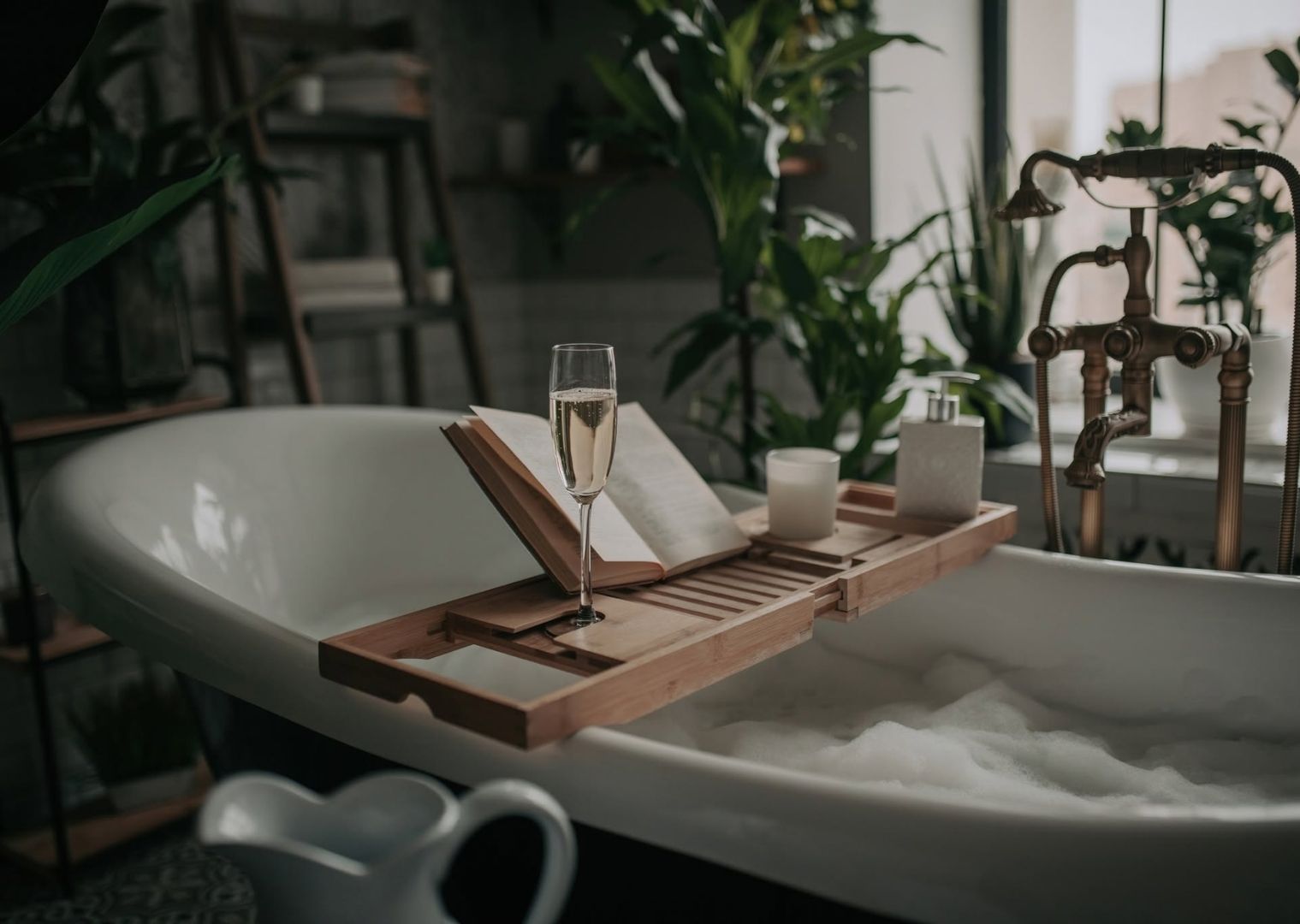 Bathtub with bubbles, wooden tray holding a glass of champagne, open book, and candle, surrounded by plants.